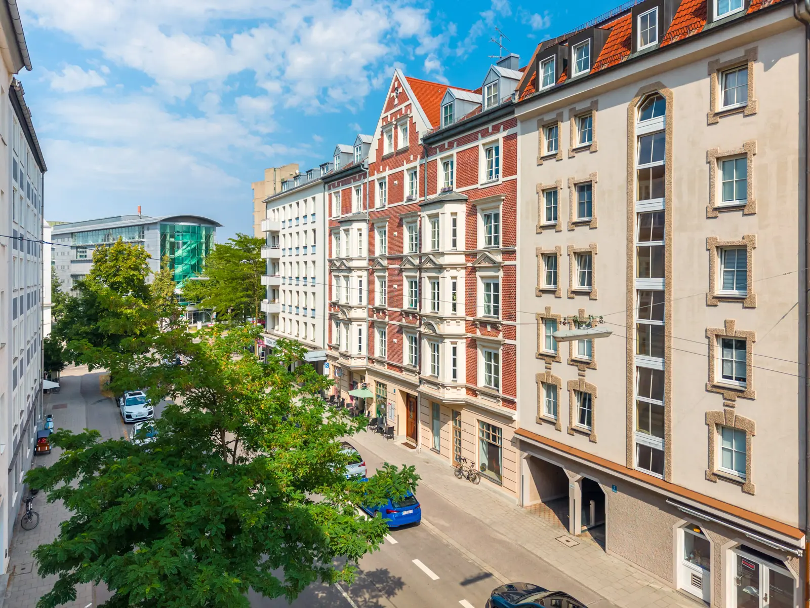 Straßenansicht von historischen und modernen mittelhohen Wohngebäuden mit am Straßenrand geparkten Autos und grünen Bäumen, die den Gehweg säumen, an einem sonnigen Tag.