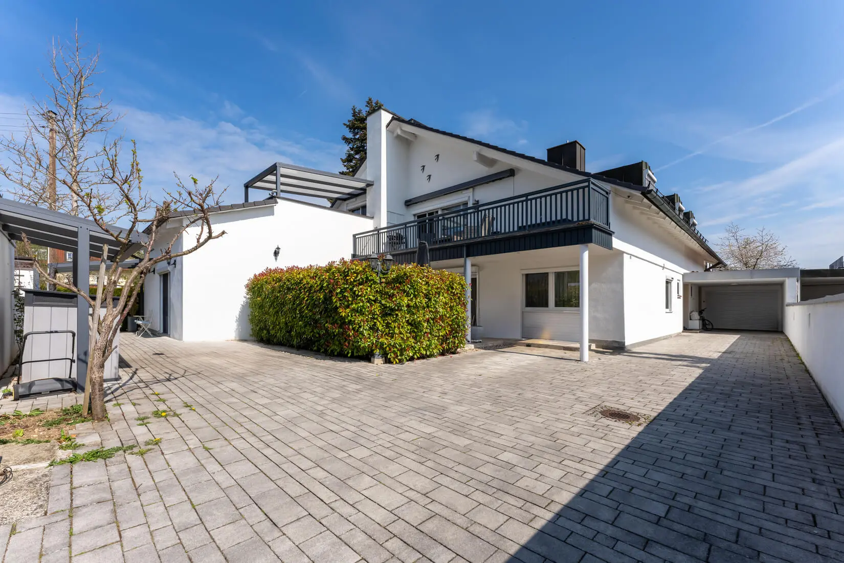 Modernes weißes zweistöckiges Haus mit Balkon, gepflasterter Auffahrt, freistehender Garage und geschnittener Hecke unter einem klaren blauen Himmel.