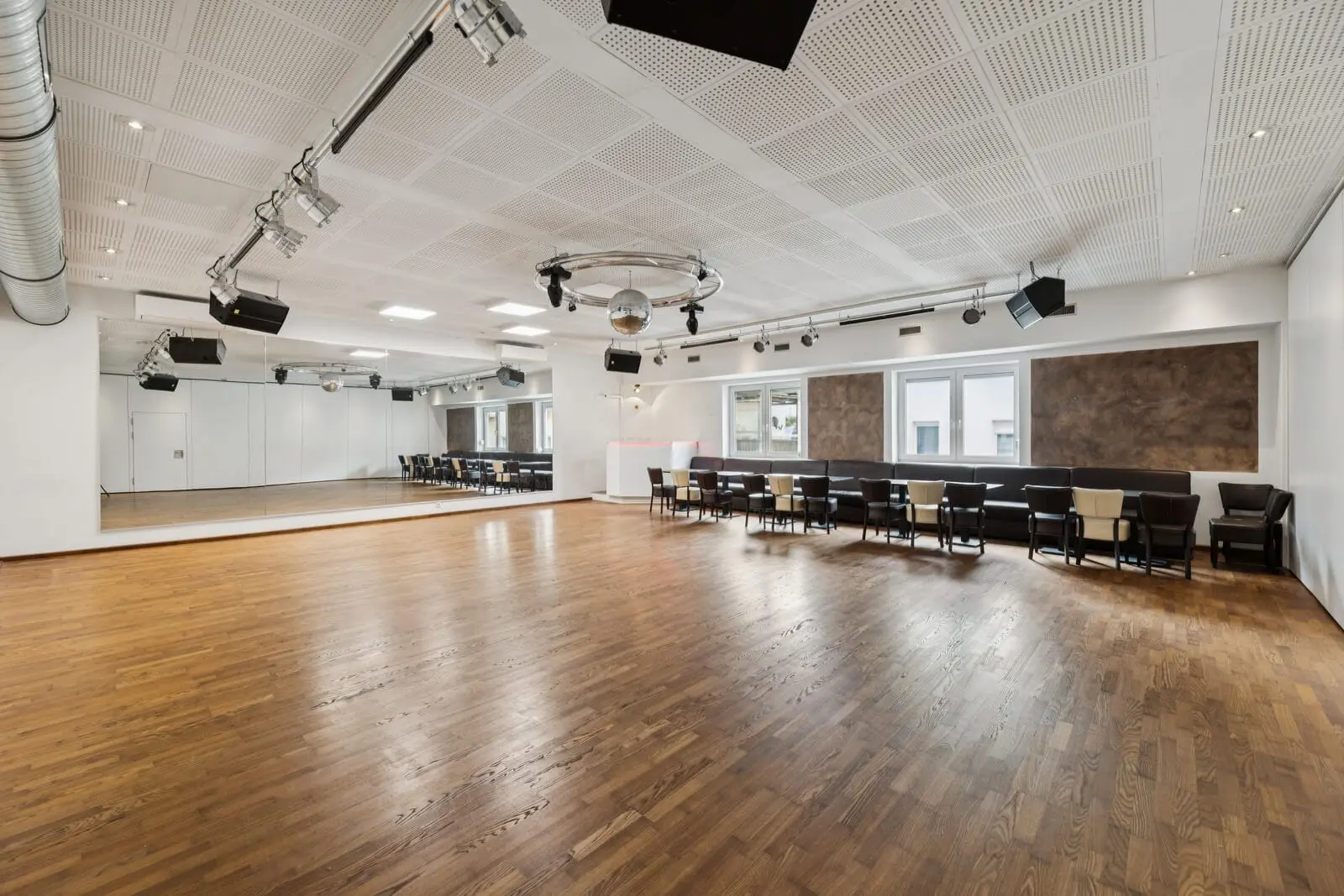 Ein geräumiges, leeres Tanzstudio mit Holzfußboden, großen Spiegeln an einer Wand, Deckenbeleuchtung, einer Discokugel und Stühlen, die an der gegenüberliegenden Wand aufgestellt sind.