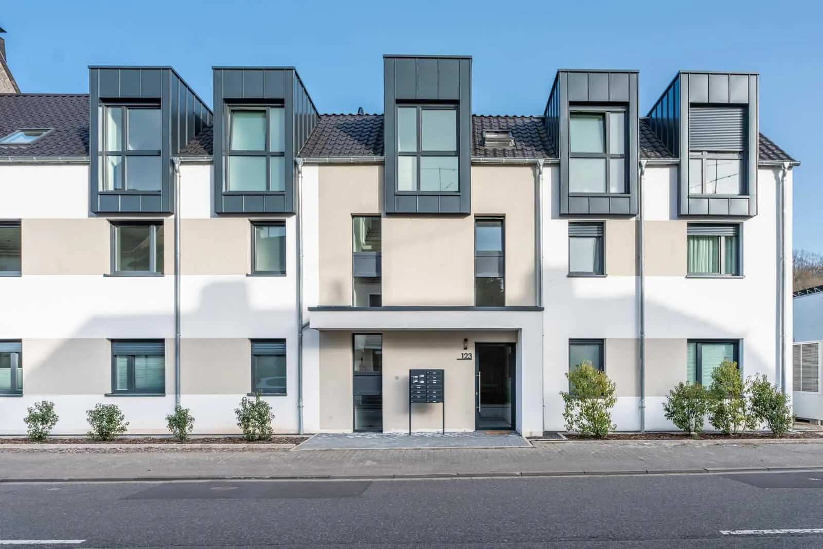 Modernes dreistöckiges Mehrfamilienhaus mit großen Kastenfenstern in den oberen Stockwerken, einer neutralen Fassade und kleinen Sträuchern vor dem Haus, das einer Straße zugewandt ist.